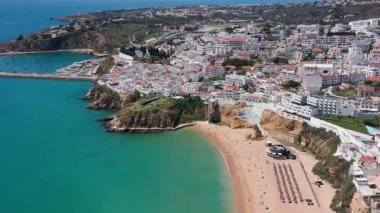 Portekiz 'in Albufeira kentindeki Praia dos Pescadores' in kumlu sahili, turkuaz denizi, uçurum kenarındaki binalar ve plaj asansörü görülebilir durumda. Sahil turizmi ve ulaşılabilir deniz kenarı yaşam tarzı kavramı