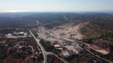 Dönemeç yolu olan sahil yamacı ocağı, hava gözlemi dağınık stoklar, vadiyi takip eden erişim yolları, uzak deniz parıltısı, kara kullanımı üzerine çevre planlayıcı perspektifi gösteriyor.