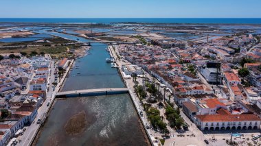 Gilao Nehri üzerindeki modern köprüyle Tavira 'nın havadan görünüşü, geleneksel binalar, tuz düzlükleri ve arka planda Atlantik Okyanusu Algarve, Portekiz' de açık mavi gökyüzünün altında.. 