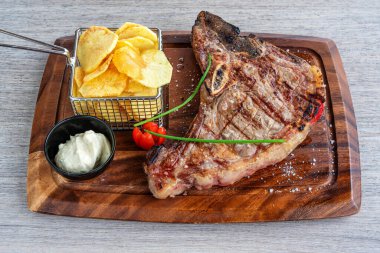 Tahta bir tahtanın üzerinde sunulan ızgara biftek yanında çıtır cipsler, taze otlar ve kremalı sosla gurme yemek deneyimini ön plana çıkarıyor.