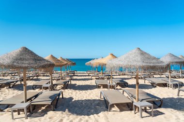 Güneş şezlongları kumlu bir sahilde sazdan şemsiyelerin altında sıra sıra dizilir. Deniz, açık mavi gökyüzü ile güneşli bir günde arka planda buluşur..