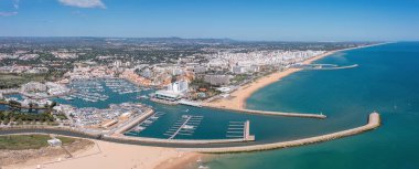 İnsanlar Portekiz 'in Algarve kentindeki Vilamoura marinasının keyfini çıkarırken, tekneler rıhtıma yanaşıyor ve sahil açık gökyüzünün altında kıyı boyunca uzanıyor. Havadan