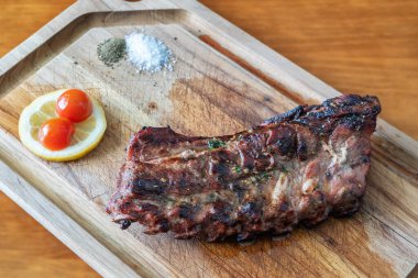 Izgara baharatlı biftek ahşap bir kesme tahtasında sunuluyor. Yanında da limon dilimleri, kiraz domatesleri ve canlı bir yemek için baharat var.