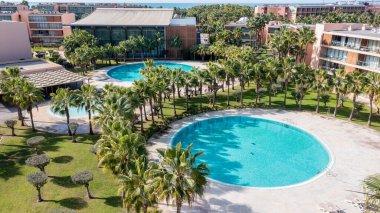 Bu hava sahnesi, güneşli bir günün ortasında dairesel yüzme havuzları, palmiye ağaçları ve yakınlarındaki binaların yer aldığı Albufeira 'nın Salgados bölgesini gösteriyor..