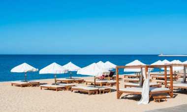 İnsanlar kumsaldaki kumların üzerinde beyaz şemsiyelerin altında uzanırlar. Güneş gökyüzünde parlıyor ve sakin dalgalar kıyıya vuruyor..