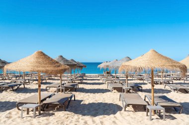 Güneş şezlongları ve saman şemsiyeleri kumlu bir sahili açık gökyüzüyle ve arka planda okyanusla dolduruyor. Birçok yer ziyaretçileri dinlenmeye ve eğlenmeye davet eder..