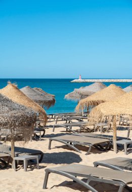 Saman şemsiyeler ve şezlonglar temiz mavi suyun yakınındaki kumlu plaja kurulur. Arka planda, açık gökyüzünün altında bir fener duruyor..