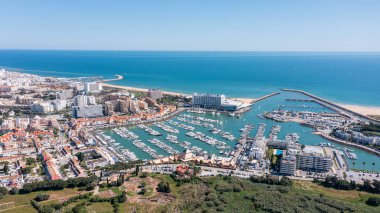 Portekiz 'in Vilamoura kentindeki marinanın keyfini çıkaran turistler, Algarve bölgesindeki açık bir gökyüzünün altında Atlantik Okyanusu manzarası ve yatları ile çevrili. Havadan