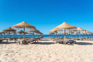 Saman şemsiyelerinin altında sakin deniz dalgaları ve arka planda açık mavi gökyüzü olan kumlu bir plajda düzenlenmiş güneşlik..