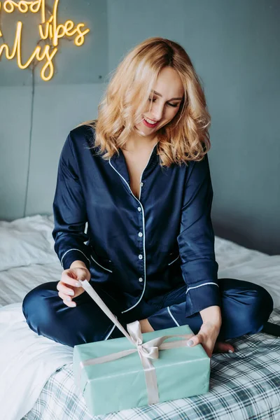 Yataktaki kız hediyeyi açıyor. Sabahleyin pijamalı bir hediyeyi açıyorum. Pijamalı kız şaşkınlıkla gülümsüyor.