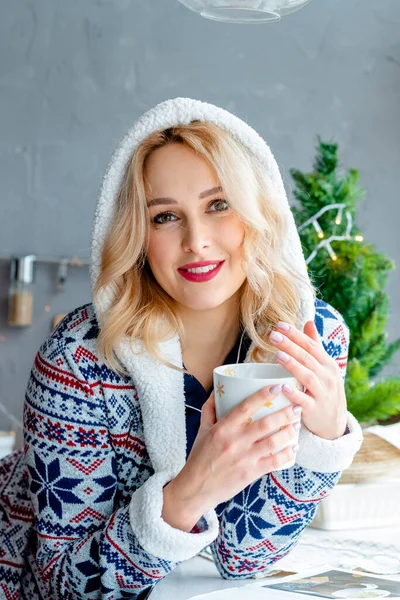 Pijamalı kız pencerenin yanında kahve içiyor. Evde pijamalı sabah kahvesi. Kız elinde bir fincanla kameraya bakıyor. Dur ve düşün. Samimi fotoğraf.