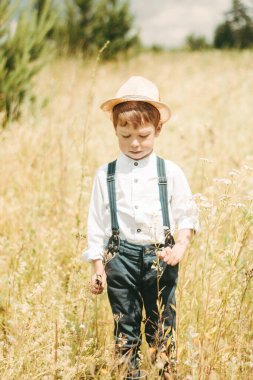 A little farmer is walking in a summer field, a little boy in a straw hat. boy in rubber boots and a white shirt. Country style