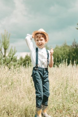 Little farmer on a summer field, happy and cute little boy in a straw hat. boy collects flowers in the field. boy in rubber boots and a white shirt. Country style