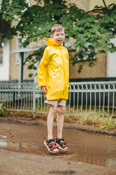 A boy in a yellow raincoat walks outside in the rain. A child jumps on the narrows. jump through muddy puddles. Happy and carefree childhood