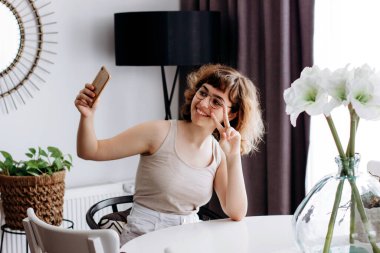 Güzel kıvırcık gülümseyen genç kız akıllı telefondan selfie çekiyor. Evde masada oturuyor.