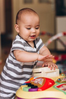 Şirin tombul bebek ayakta duruyor ve çocukların müzik sehpasıyla oynuyor. Her yerde bir sürü renkli oyuncak var..