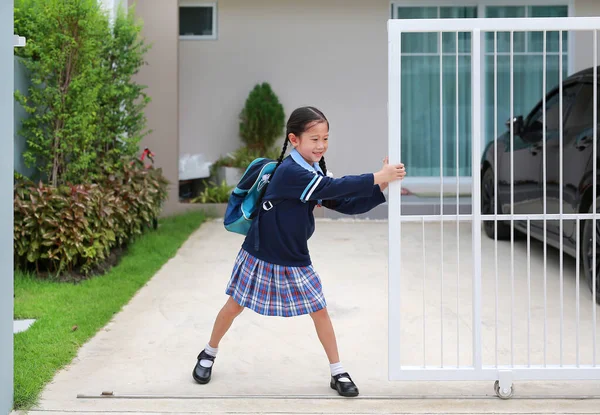Cheerful asian little child girl in kindergarten uniform trying push and pull door fence of house to close or open before leave home to go to school at morning.