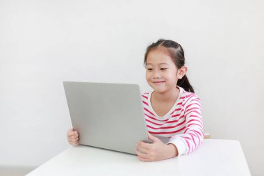 Masada oturup dizüstü bilgisayar kullanan mutlu küçük Asyalı kız. Evde kal.