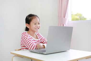 Küçük mutlu Asyalı kız masada oturuyor ve dizüstü bilgisayar kullanıyor.