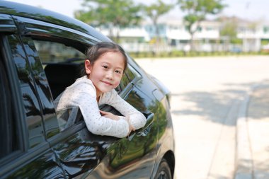 Arabada oturmuş, arabanın camından kameraya bakarak gülümseyen Asyalı küçük kız çocuğu.