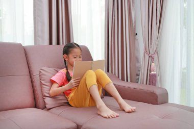 Mutlu Asyalı küçük kız kitap okuyor. Evdeki kanepede yatıyor.