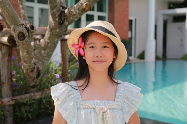 Pembe Plumeria çiçeğiyle süslenmiş hasır şapkalı Asyalı bir kız ve açık mavi beyaz desenli dantelli bir elbise. Yüzme havuzu ve yemyeşil bir arka planı var..