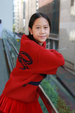 Neşeli Asyalı kız kameraya gülümserken yürüyen merdivenin çelik ve cam parmaklıklarına yaslanırken parlak kırmızı örgü süveter giyip kırmızı pileli etek giyiyor..