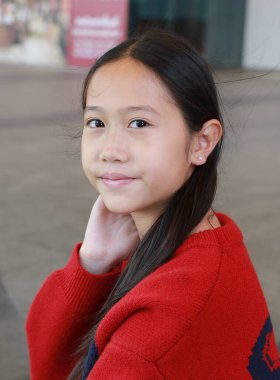 Uzun siyah saçlı güzel bir Asyalı kız, rahat kırmızı örgü süveter giyiyor, eli hafifçe boynunun arkasında dinlenirken dışarıda poz verirken nazikçe gülümsüyor..