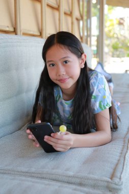 Hoş, genç Asyalı bir kız rahat bir kanepede uzanmış elinde akıllı bir telefon ve elinde sarı bir lolipop tutarken mutlu bir şekilde gülümsüyor..