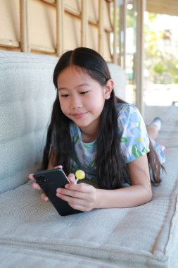 Neşeli bir Asyalı genç kız rahat bir kanepede uzanıyor, akıllı telefonunu kullanarak tatlı sarı bir lolipopun tadını çıkarıyor..