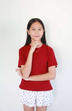 Kırmızı tişörtlü ve benekli şortlu çekici bir Asyalı kız eli çenesinde ve beyaz arka planda soyutlanmış nazik bir gülümsemeyle düşünceli bir poz veriyor..