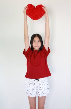 Kırmızı tişörtlü neşeli Asyalı kız iki eliyle kırmızı kalp şeklindeki yastığı kaldırıp beyaz arka planda duruyor..