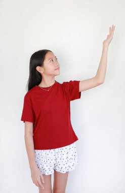 Kırmızı tişörtlü ve benekli etekli genç Asyalı kızın portresi beyaz arka planda duruyor, gülümsüyor ve elini kaldırıyor ya da bir şey gösteriyor..