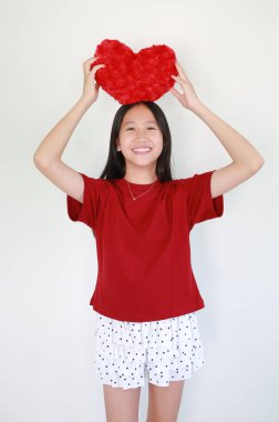 Kırmızı tişörtlü ve benekli etekli neşeli Asyalı kız kırmızı kalp şeklindeki pelüş yastığı beyaz arka planda tutarken ışıl ışıl gülümsüyor..