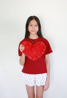 Kırmızı tişört ve beyaz puantiyeli şort giyen neşeli genç Asyalı kız, ayağa kalkar ve yumuşak bir gülümsemeyle yumuşak kırmızı kalp şeklinde bir yastık tutar..