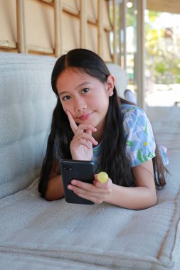 Hoş, genç Asyalı bir kız rahat bir kanepede uzanmış elinde akıllı bir telefon ve elinde sarı bir lolipop tutarken mutlu bir şekilde gülümsüyor..