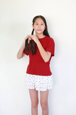 Kırmızı tişörtlü ve puantiyeli tatlı Asyalı kız uzun siyah saçlarını beyaz arka planda tutarken ve örerken hafifçe gülümsüyor..