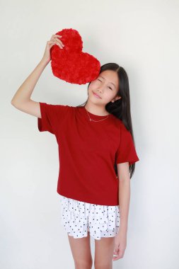 Kırmızı tişört giyen genç Asyalı kızın portresi, beyaz arka planda başının yanında kırmızı bir yastık tutarken ayakta duruyor ve göz kırpıyor..