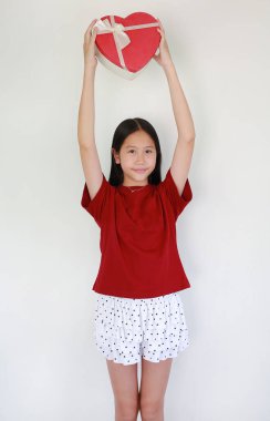 Kırmızı tişörtlü ve benekli etekli genç Asyalı kızın portresi kafasının üstünde kalp şeklinde bir hediye kutusu taşıyor ve beyaz arka planda kameraya gülümsüyor..