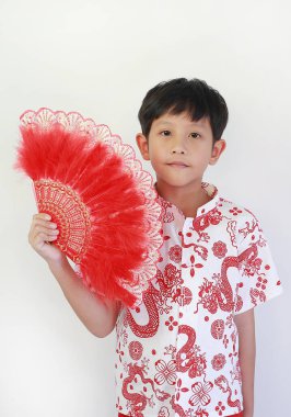 Beyaz ejderha desenli tişörtlü yakışıklı Asyalı çocuk Çin Yeni Yılı 'nı kutluyor, yumuşak bir gülümsemeyle poz veriyor ve elinde dekoratif kırmızı pervane tutuyor..