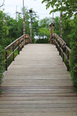 Asılı ahşap etiketlerle süslenmiş, yemyeşil ağaçlara doğru uzanan uzun tahta bir köprünün huzurlu manzarası..