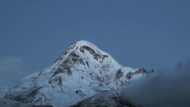 Stepantsminda, Gergeti, Georgië. Mount Kazbek bedekt sneeuw in de winter zonsopgang. Morning Dawn gekleurde top van de berg in roze-oranje kleuren. Georgisch natuurlandschap. Tijdsverloop, Timelapse, Tijdsverloop — Stockvideo