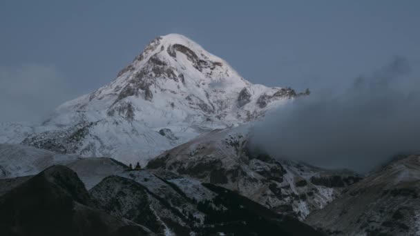 Stepantsminda, Gergeti, Georgia. Mount Kazbek zakryl sníh v zimním východu slunce. Ranní úsvit barevný vrchol hory v růžových barvách. Úžasná zimní gruzínská přírodní krajina. Časová prodleva 4K — Stock video