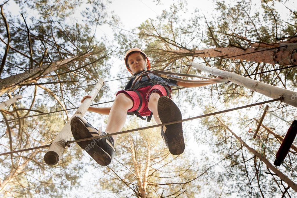 A boy wearing a safety harness and a helmet walking in a rope park.