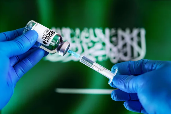 Doctor preparing vial of vaccine injection for the vaccination plan against diseases in Saudi Arabia. Injecting dose of vaccine in syringe for infections prevention in front of the Saudi Arabia flag