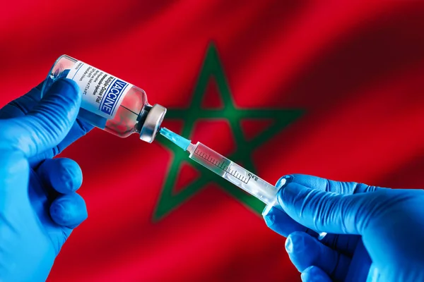 Doctor preparing vial of vaccine injection for the vaccination plan against diseases in Morocco. Injecting dose of vaccine in syringe for infections prevention in front of the Morocco flag