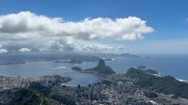Corcovado 'dan Rio de Janeiro' nun panoramik görüntüsü, Sugarloaf Dağı, Guanabara Körfezi, Santos Dumont Havalimanı ve Atlantik kıyı şeridi parlak, parçalı bulutlu hava altında.