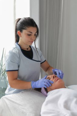 Üniformalı genç bir estetisyen, temiz bir spa ortamında olgun bir bayan müşteriye profesyonel yüz bakımı yapıyor..