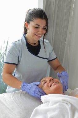 Üniformalı genç bir estetisyen, temiz bir spa ortamında olgun bir bayan müşteriye profesyonel yüz bakımı yapıyor..