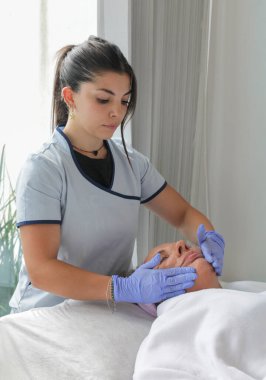 Üniformalı genç bir estetisyen, temiz bir spa ortamında olgun bir bayan müşteriye profesyonel yüz bakımı yapıyor..